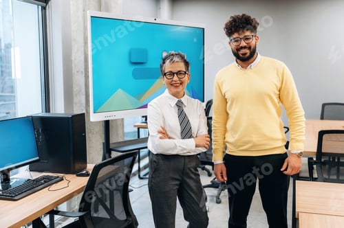 Preview: Business professionals standing confidently in modern office space with computers and digital screen