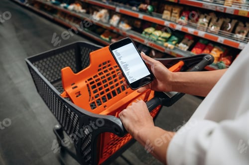 Preview: Person holding smartphone while pushing shopping cart in grocery store aisle, surrounded by shelves