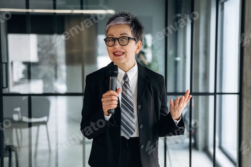 Preview: Businesswoman with short gray hair in a black suit and tie holds a microphone while speaking in a mo