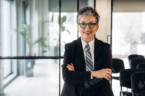 Preview: Businesswoman with short gray hair wearing a black suit and glasses stands confidently with arms cro