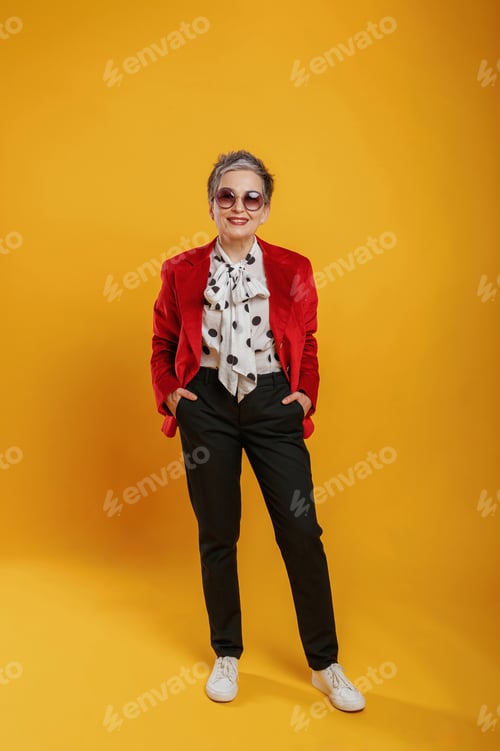 Preview: Stylish elderly woman in vibrant red blazer and polka dot blouse poses confidently against a bright