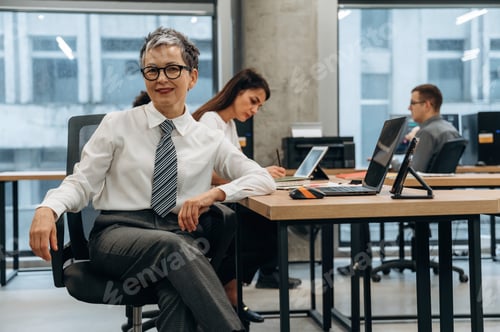 Preview: Confident businesswoman in modern office setting, seated with colleagues working at desks, showcasin
