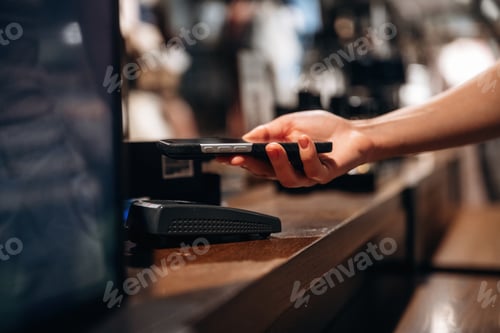 Preview: Hand holding smartphone near contactless payment terminal on wooden counter in modern retail store w
