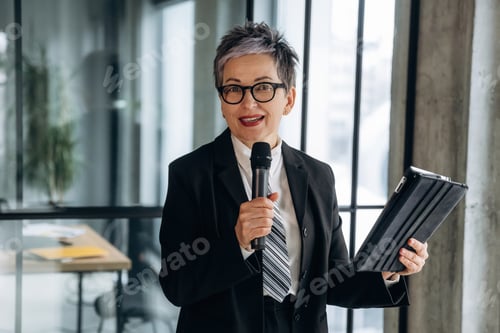 Preview: Businesswoman with short gray hair wearing glasses holds a microphone and tablet while standing in a