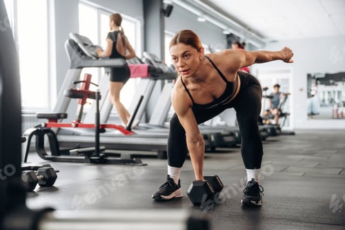 Preview: Female fitness enthusiast performing bent-over dumbbell row exercise in modern gym with various work