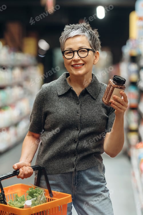 Preview: Senior woman with short gray hair wearing glasses holds a spice jar and shopping basket while standi