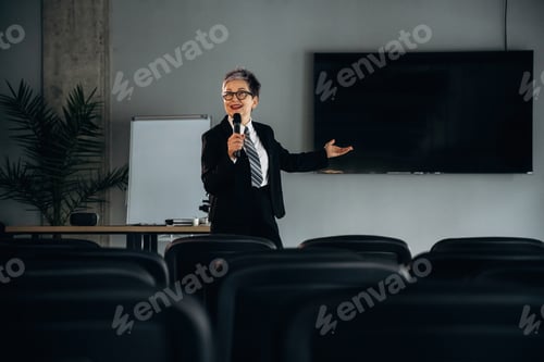 Preview: Businesswoman presenting in a modern office setting, holding a microphone, with a large screen and e