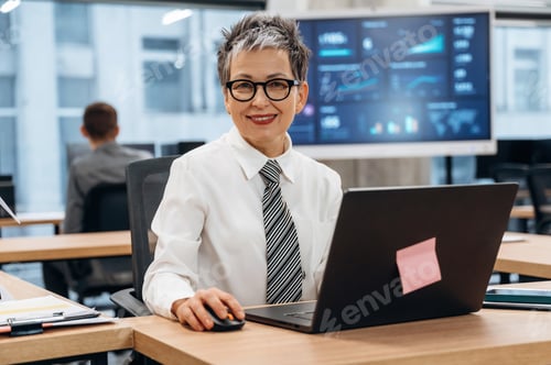 Preview: Businesswoman with short gray hair is smiling while working on laptop in modern office, showcasing a