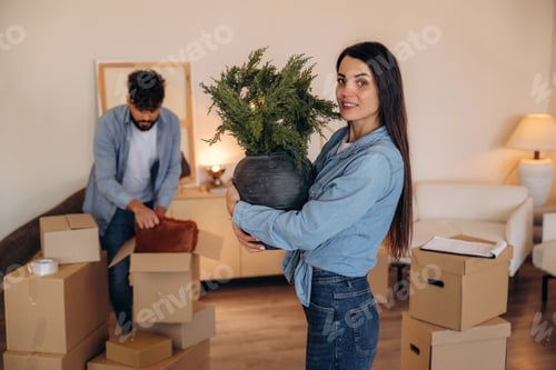 Preview: Looking at the camera and smiling. Positive couple placing a potted plant in their new apartment dur