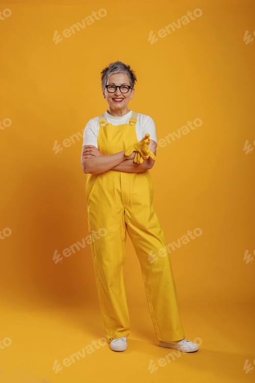 Preview: Senior woman wearing yellow overalls and glasses is smiling confidently with arms crossed, showcasin