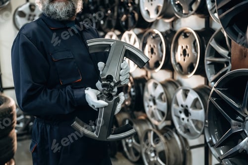 Preview: Caucasian male technician in blue coveralls holds a car wheel rim in front of a display of various a