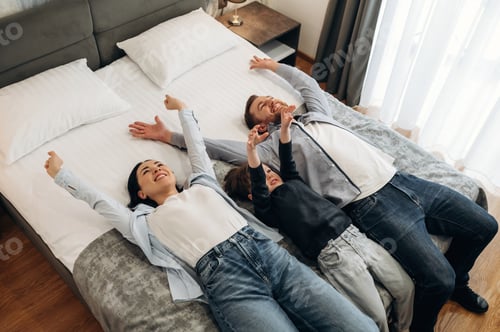 Preview: Family of three lying on a hotel bed, smiling and enjoying their time together with arms raised, bri