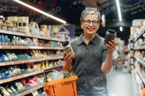 Preview: Senior woman with short gray hair wearing glasses holds jars of spices while shopping in a grocery s
