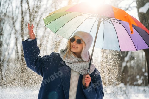 Preview: Young woman in winter attire holding a colorful umbrella while enjoying snowfall in a forest, sunlig