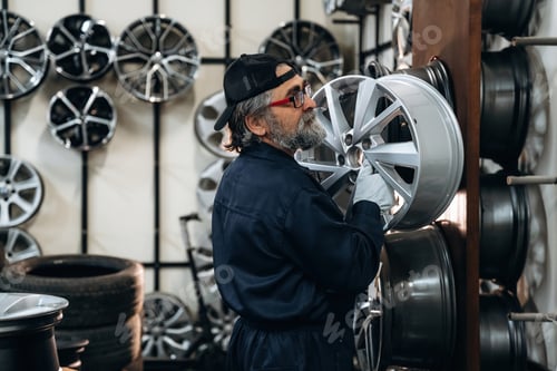 Preview: Male technician in dark coveralls inspects a silver car wheel in an automotive shop with multiple wh