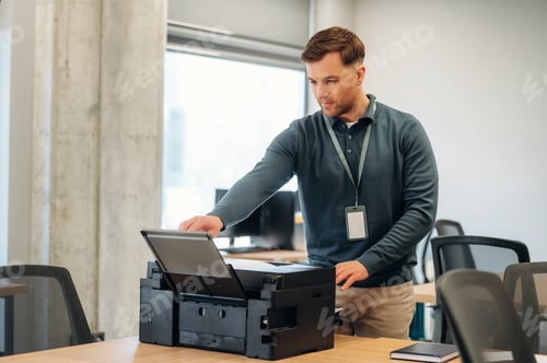 Preview: Male office worker in blue sweater operates a printer on a wooden desk in a modern workspace with co