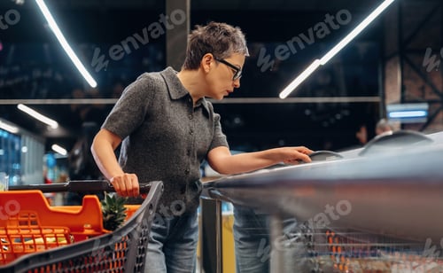 Preview: Short-haired woman wearing glasses inspects items on a shelf while pushing a shopping cart in a mode