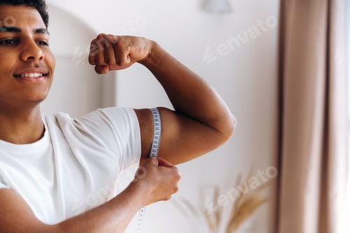 Preview: Young male measuring bicep size with tape measure while smiling in a bright indoor setting with soft