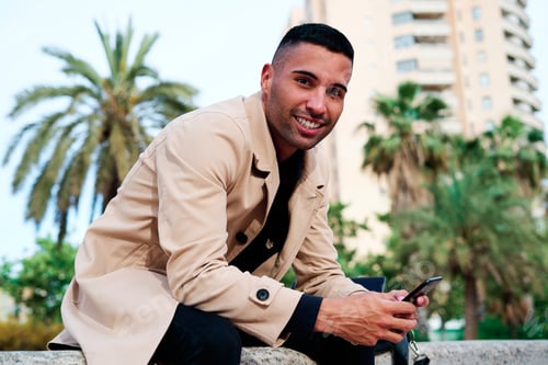 Preview: Young man in a light coat using smartphone while sitting outdoors in a park with palm trees in the b