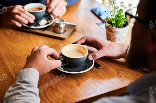 Preview: Man holding cappuccino at cafe table while another person prepares coffee in background