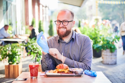 Preview: Bearded man putting on glove, eating burger at outdoor cafe, sitting at table with fries and drink