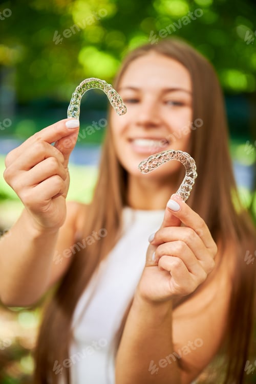 Preview: Young woman holds clear aligners and smiles at camera. Invisible orthodontic trays outdoors
