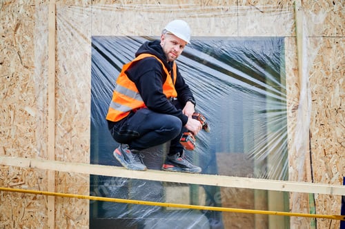 Preview: Construction worker in helmet and safety vest pausing with power drill beside timber facade