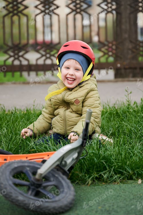 Preview: Smiling toddler in helmet sitting on grass near fallen balance bike in park. Happy child after ride