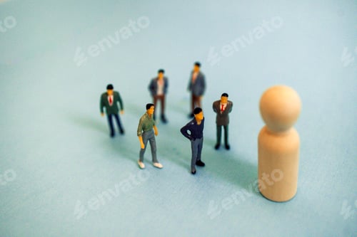 Preview: Group of miniature figurines in business attire standing around a larger wooden figure on a light bl