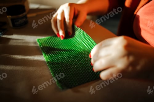 Preview: Female artisan with red nails carefully folds green beeswax sheet for candle making on a beige surfa