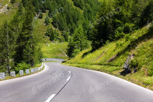 Preview: Scenic winding alpine road curves green meadow forested slopes mountains clear sky. Scenic countrysi