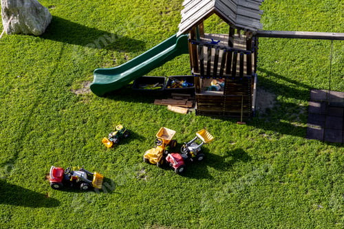Preview: Wooden playground green slide scattered toy tractors bright grass sunlight calm outdoor atmosphere.