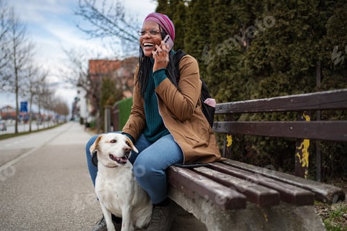 Preview: Happy woman talking on phone with dog at park bench