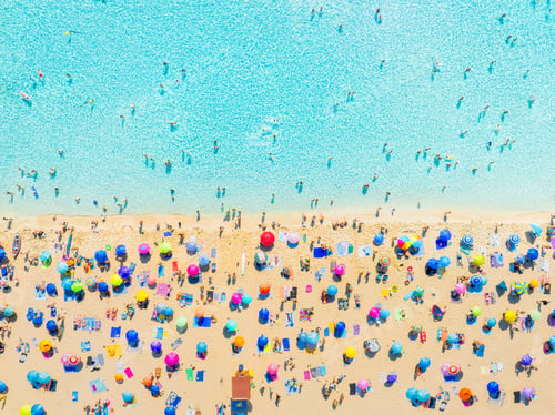 Preview: Aerial drone view of a crowded Mediterranean beach with people and colorful umbrellas.