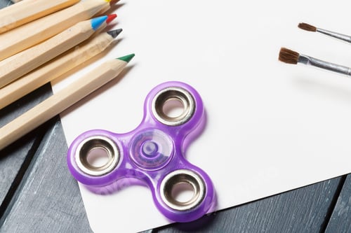 Preview: Purple fidget spinner rests on a blank sheet next to colored pencils and brushes
