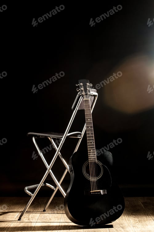 Preview: Black guitar rests against a silver stool in a dimly lit room with wooden floor