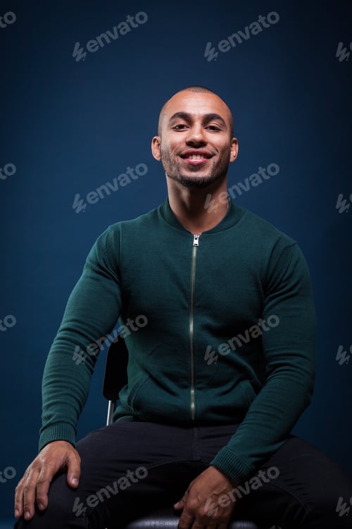 Preview: Man sitting on chair in front of blue background smiling confidently during studio session