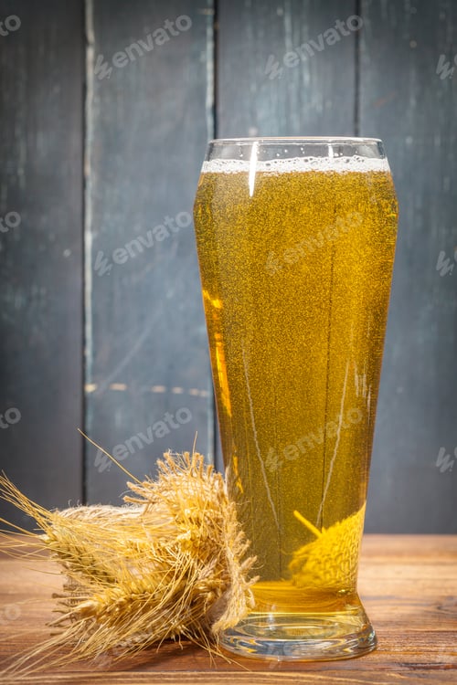 Preview: Golden beer sits in a tall glass next to barley on a wooden table in a rustic setting
