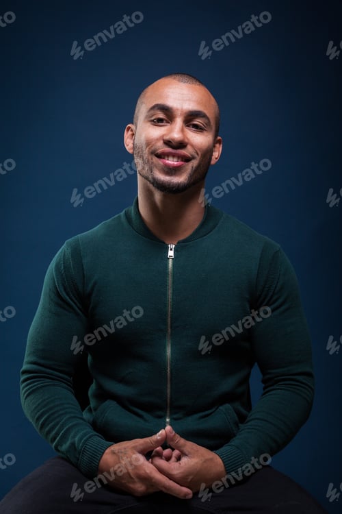 Preview: Man sitting with smile, wearing green sweater, against blue background in indoor setting