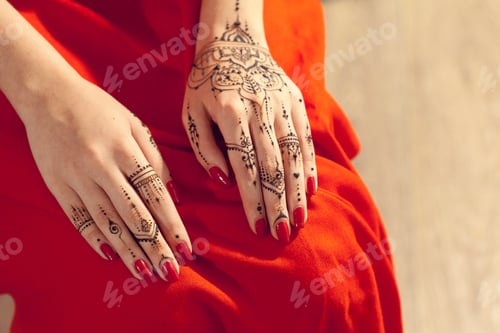 Preview: Hands with red nails and henna design sitting on a red fabric surface