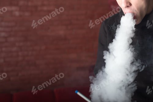 Preview: Person exhales smoke from a vape device in a dimly lit indoor space during the evening hours