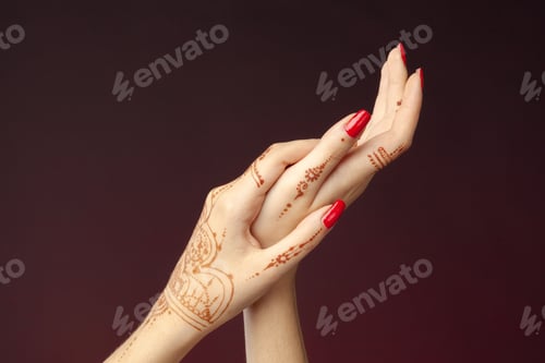 Preview: Hands with henna design hold each other and show bright red nails in soft lighting