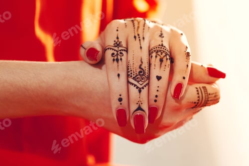 Preview: Hands with red nails and henna design holding each other indoors in natural light