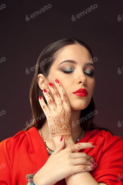 Preview: Woman with henna and bright nail polish poses with hand on face in studio setting