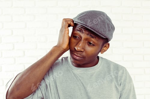Preview: Young man with hat looks thoughtful while holding his head in a room with white walls