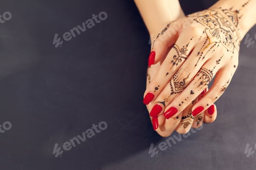 Preview: Hands with henna design and red nails resting together on a dark surface