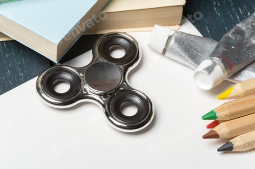 Preview: Fidget spinner rests on a blank paper with art supplies and books nearby