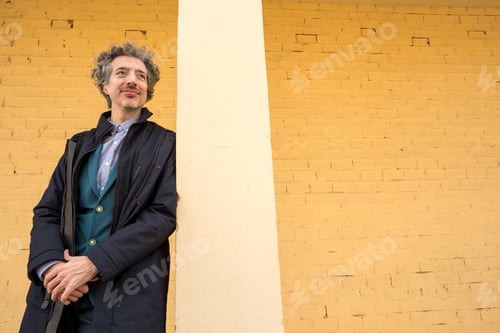 Preview: Confident man with graying hair standing against a yellow brick wall, looking away with a thoughtful