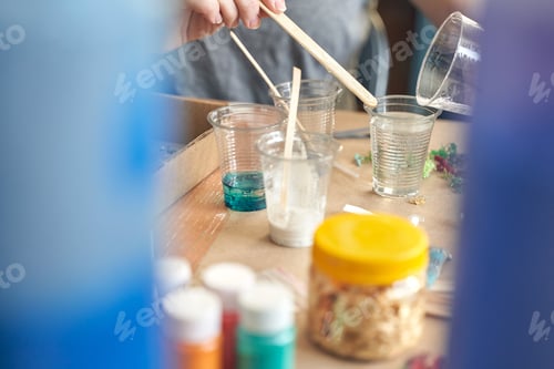 Preview: Hands pouring resin into clear cups for crafting project on worktable with paint