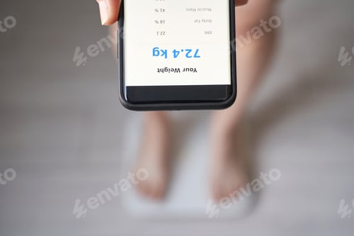 Preview: Overhead Close-Up Of Smartphone Held Above Blurred Feet On A Scale Showing 72.4 Kg For Fitness App